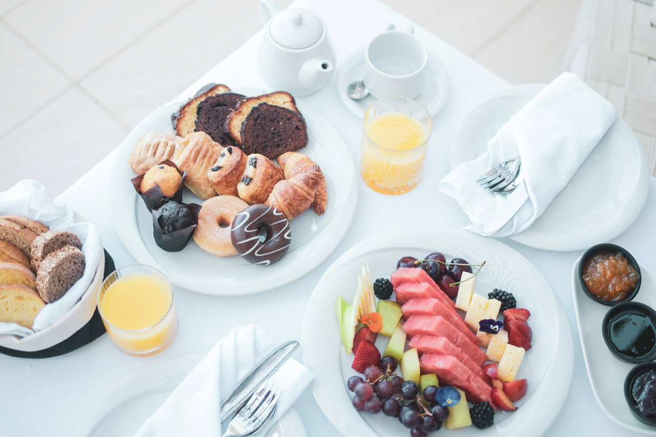 Gedeckter Frühstückstisch von oben mit Gebäckteller, Obstplatte, Brotkorb, Orangensaft, Teekanne, Marmeladen und weißen Servietten