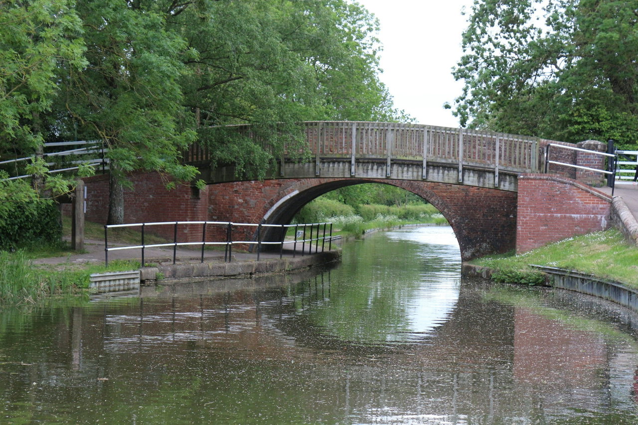 Alte Bogenbrücke aus Backstein mit Holzgeländer über einen schmalen, von Bäumen gesäumten Kanal.