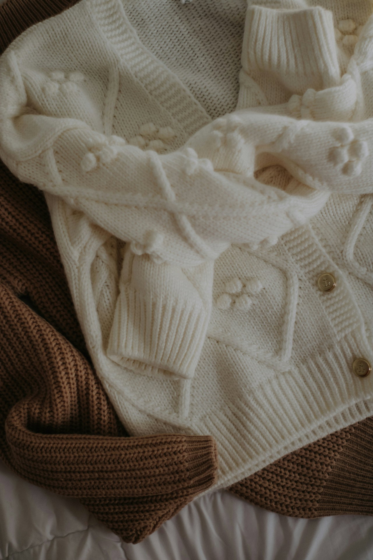 Cremefarbener Grobstrick-Cardigan mit Knöpfen, flach gelegt neben einem braunen Strickteil