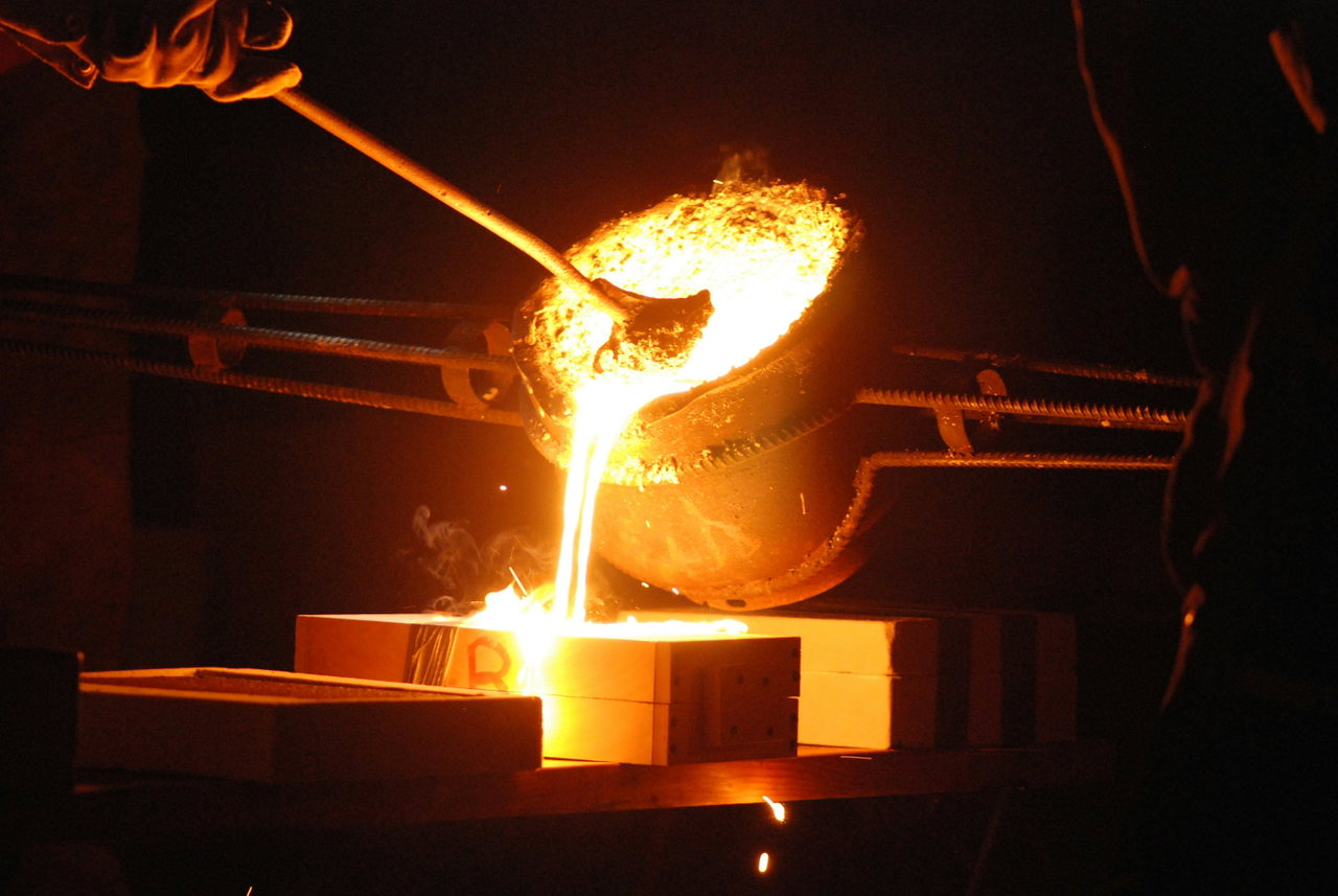 Flüssiges Metall fließt aus einer Gießkelle in eine Form, Funken und warmes Licht in der Gießerei