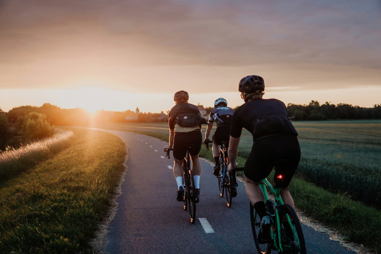 Drei Rennradfahrer fahren im Gegenlicht der tief stehenden Sonne auf einer schmalen, kurvigen Landstraße