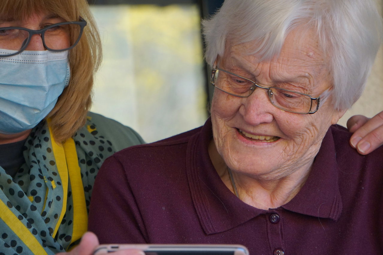 Older woman smiling as she looks at a device, with a care worker in a face mask beside her