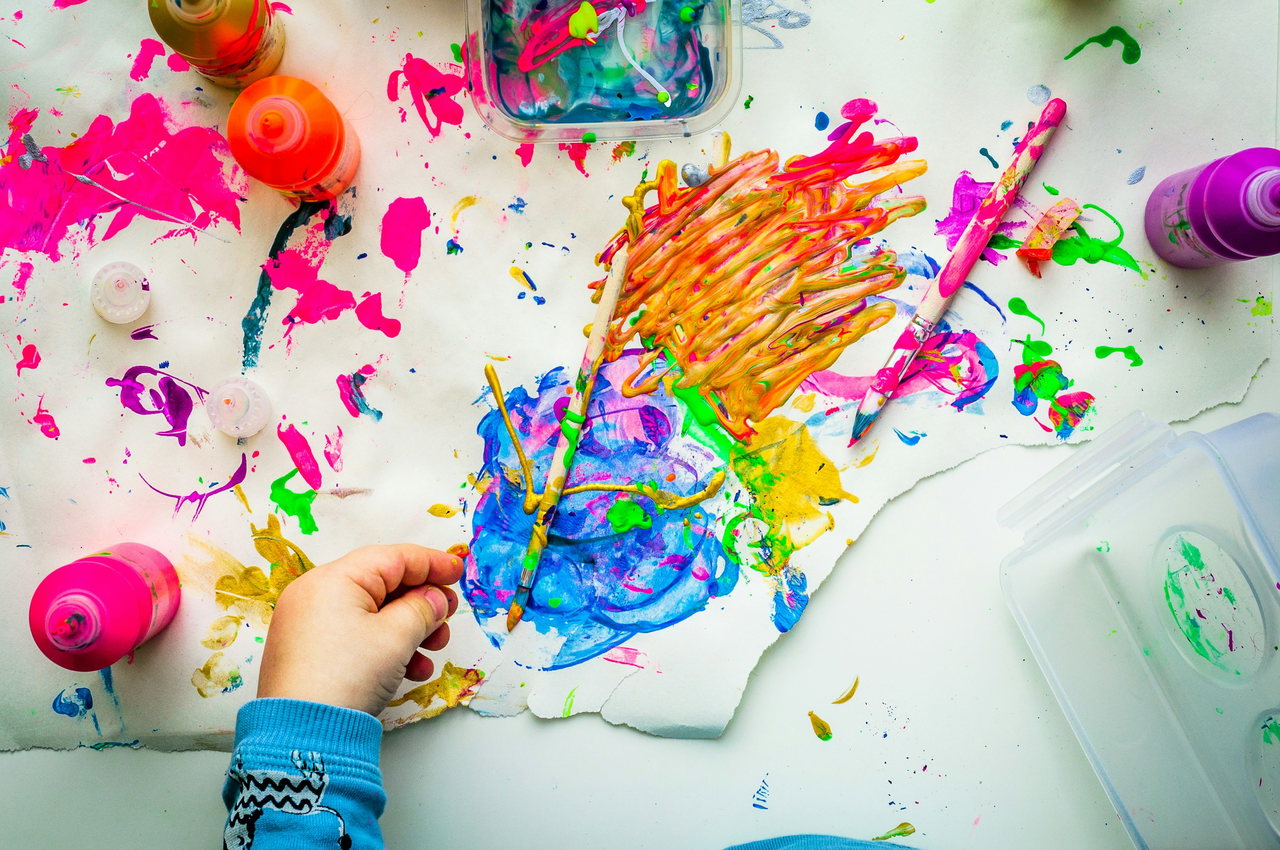 Kinderhand malt auf weißem Papier mit bunten Fingerfarben ein farbenfrohes Bild, Farbtöpfe und Farbspritzer ringsum