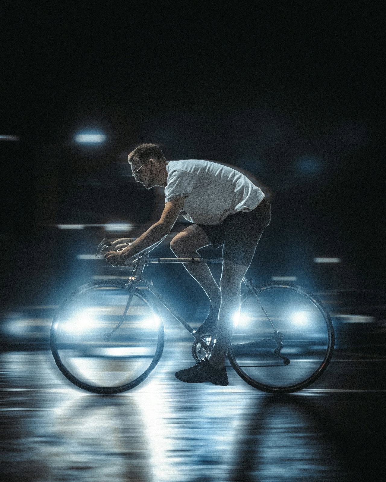 Radfahrer im weißen Shirt fährt nachts auf einem Rennrad über nasse Straße, die Laufräder leuchten hell auf