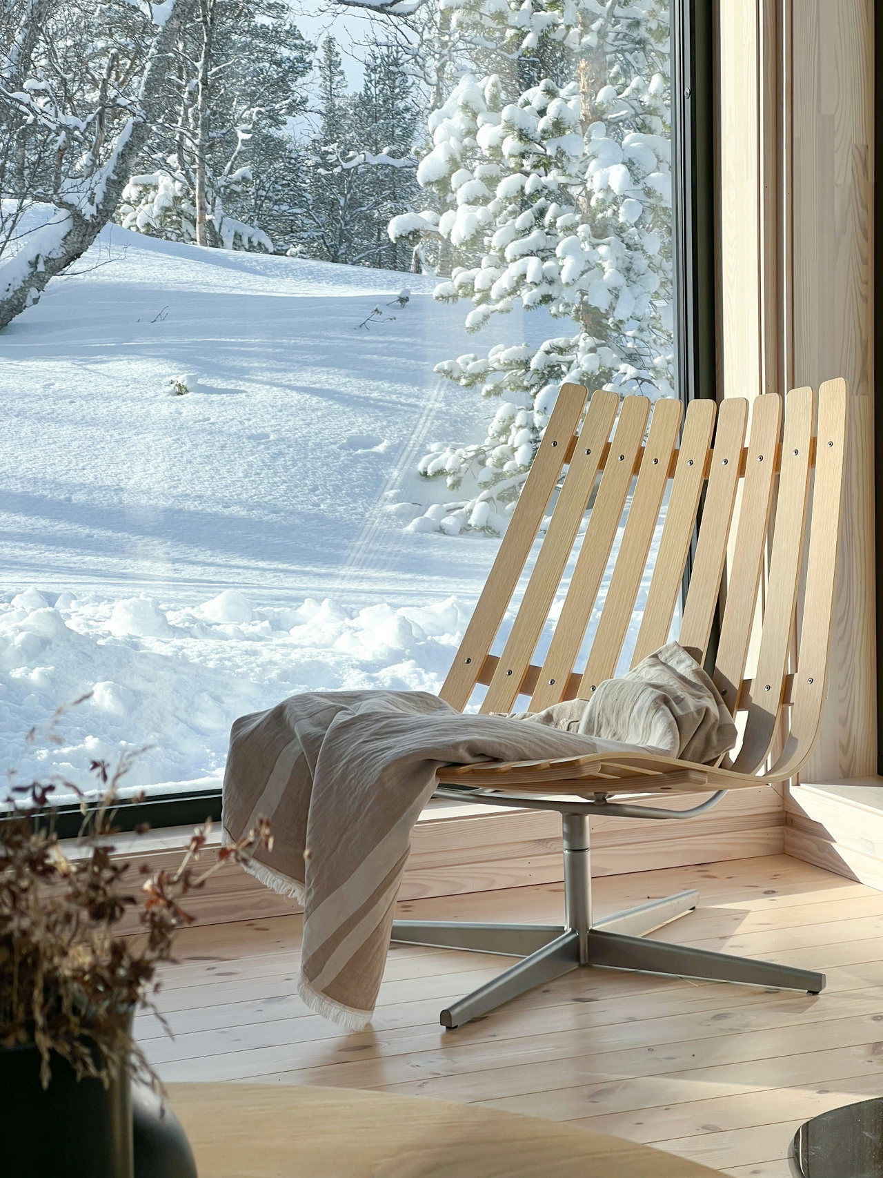 Heller Holzsessel mit Wolldecke am großen Fenster, draußen verschneite Bäume im Sonnenlicht