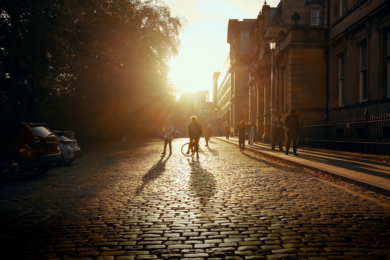 Radfahrer und Fußgänger als Silhouetten auf einer Kopfsteinpflasterstraße zwischen alten Steinhäusern im Gegenlicht der Abendsonne.