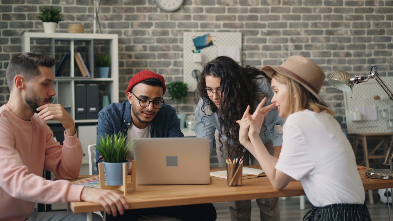 Vier junge Kreative diskutieren am Tisch vor einer Ziegelwand, einer arbeitet am Laptop