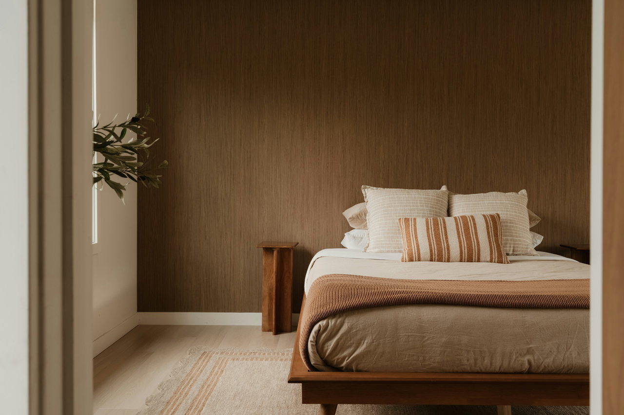 Schlafzimmer mit niedrigem Holzbett, hellen Kissen und gestreiftem Zierkissen vor einer Wand mit dunkler Holzstruktur, Blick durch die geöffnete Tür.
