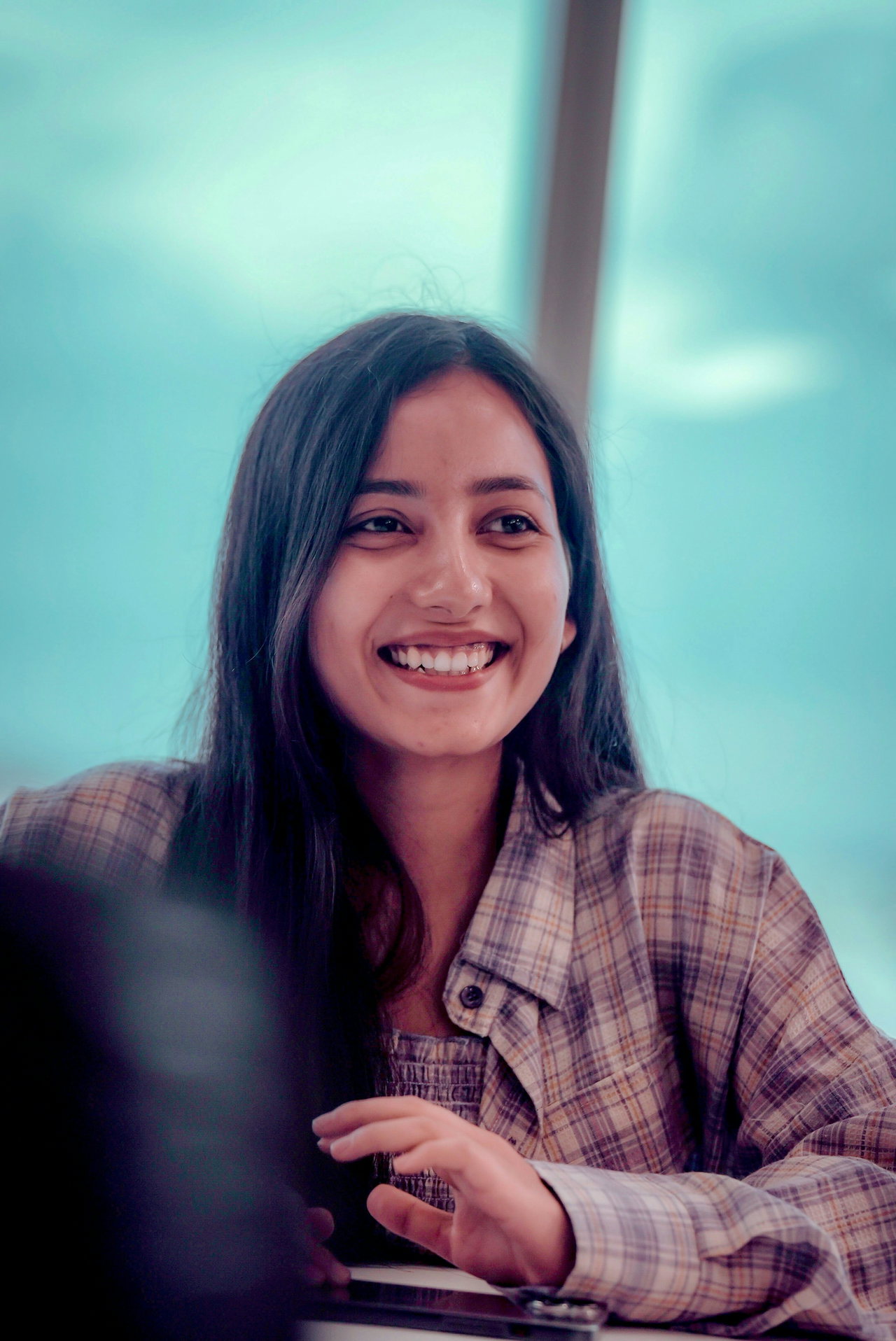 Laughing young woman with long dark hair and a checked shirt against a blurred turquoise background