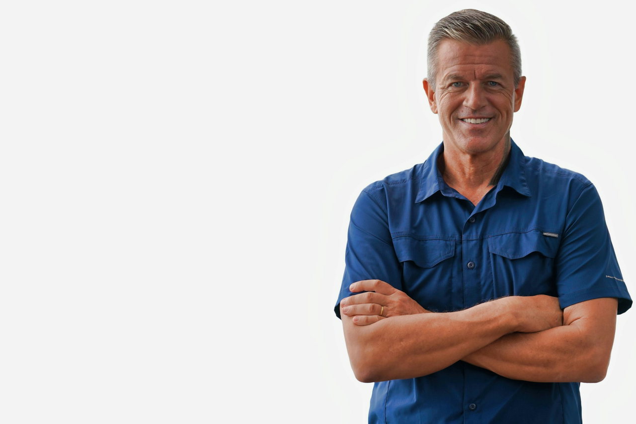 Mann mit grauen Haaren in blauem Arbeitshemd steht mit verschränkten Armen lächelnd vor hellem Hintergrund