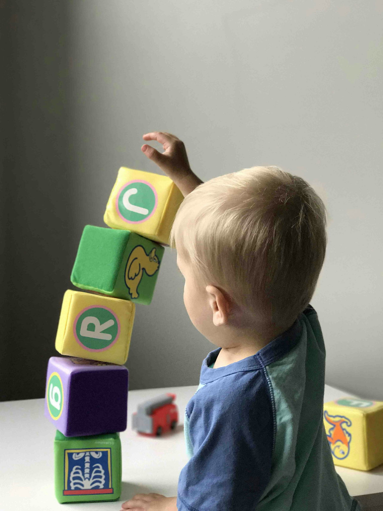 Kleinkind in blauem Shirt stapelt bunte Bauklötze mit Buchstaben zu einem hohen Turm, von der Seite fotografiert