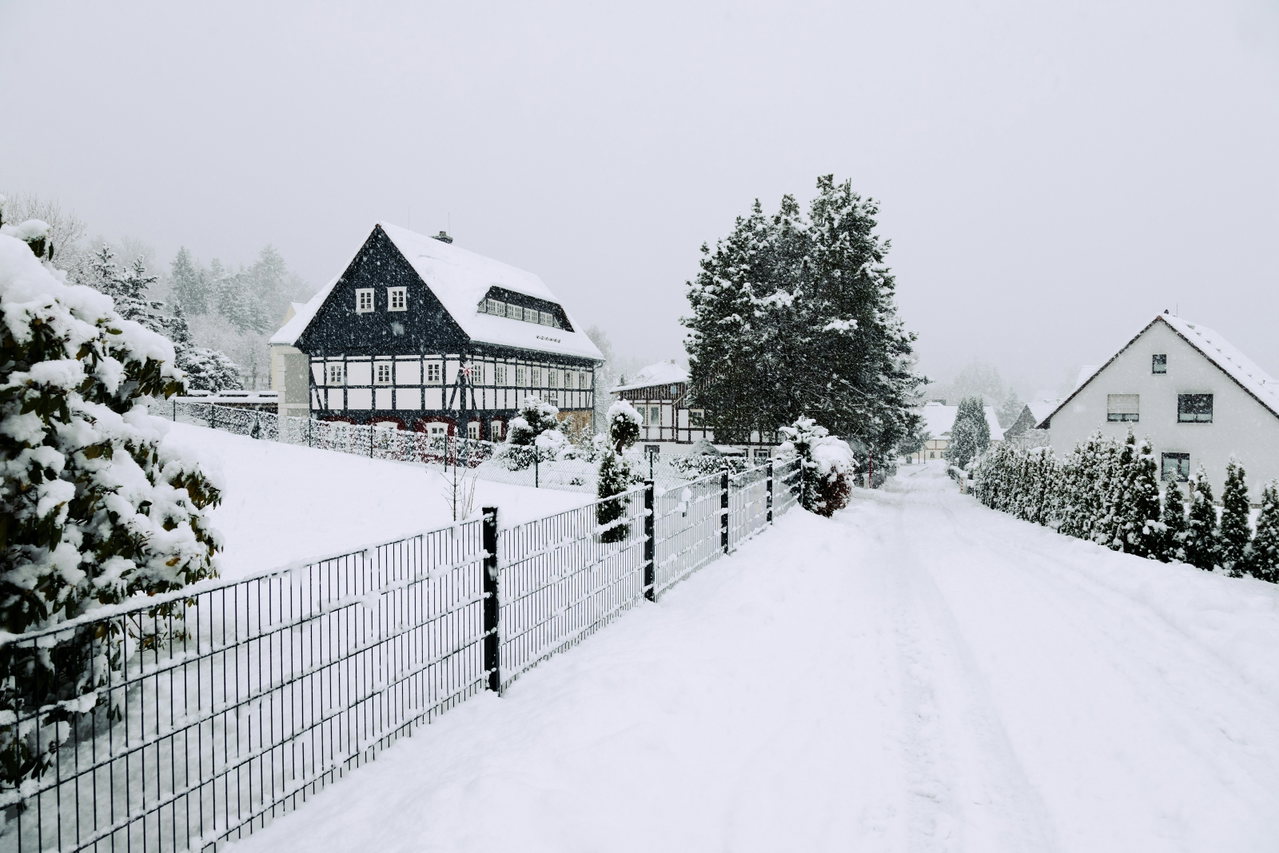 Verschneite Dorfstraße mit schwarz-weißem Fachwerkgasthof und Gartenzaun unter ruhigem Winterhimmel
