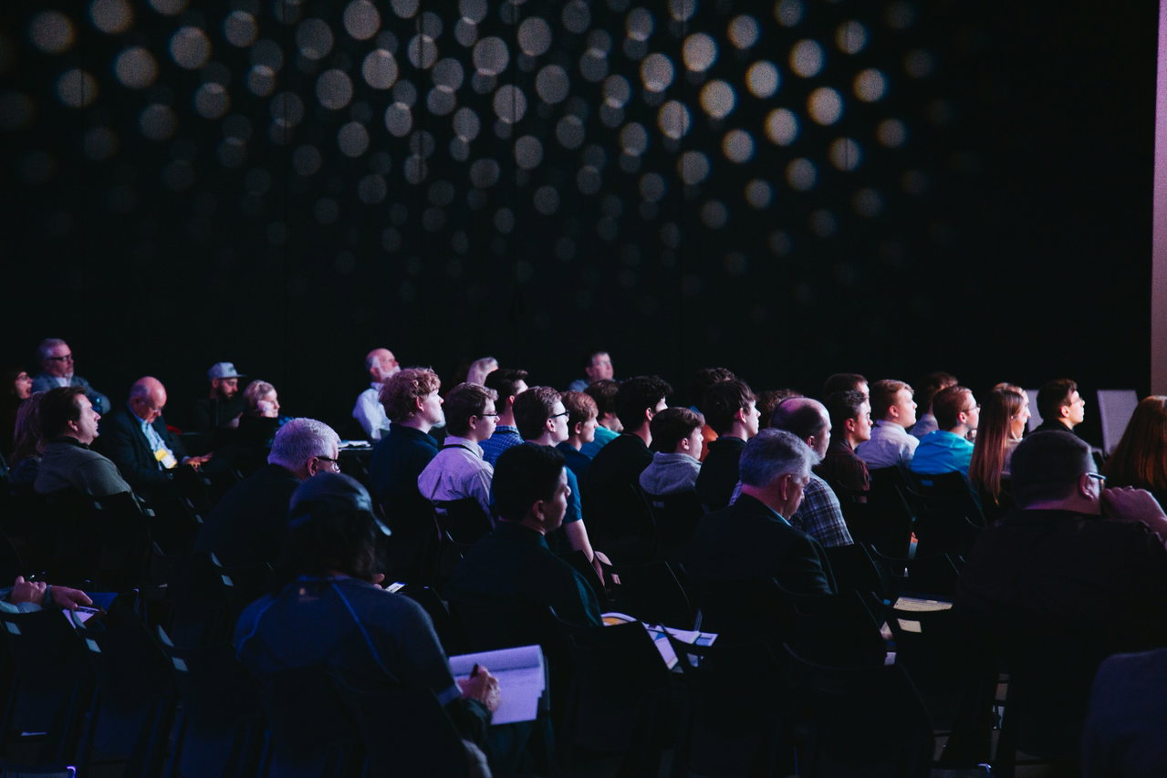 Konferenzpublikum von hinten im abgedunkelten Saal, einige Teilnehmende machen sich Notizen