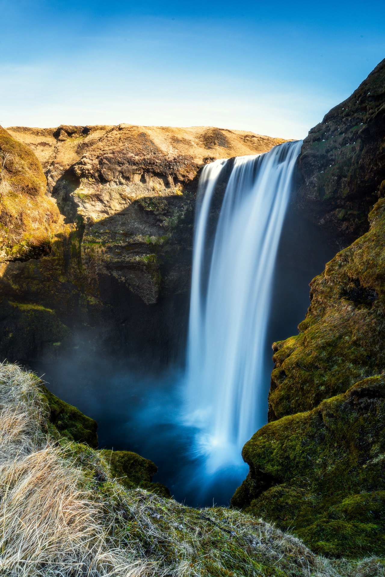 Ein hoher Wasserfall stürzt unter blauem Himmel an moosgrünen Felsen hinab