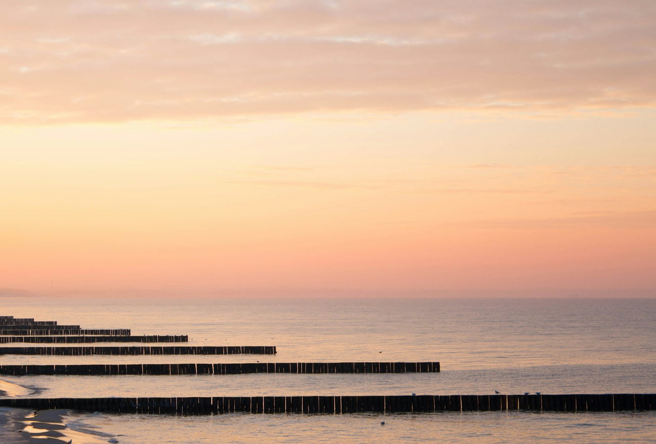 Pastellfarbener Sonnenuntergang über der ruhigen Ostsee mit langen hölzernen Buhnen, die ins Wasser ragen