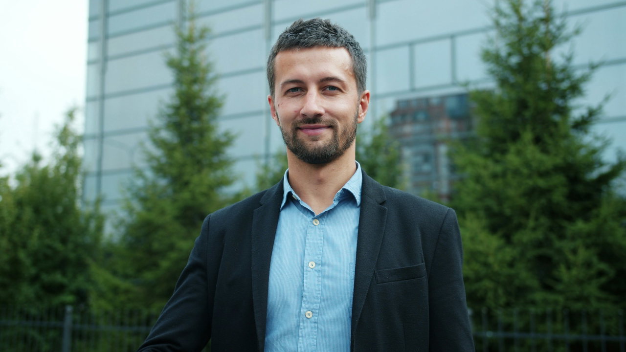 Bärtiger Mann mit dunklem Sakko über hellblauem Hemd ohne Krawatte, freundlich in die Kamera blickend, grüner Hintergrund
