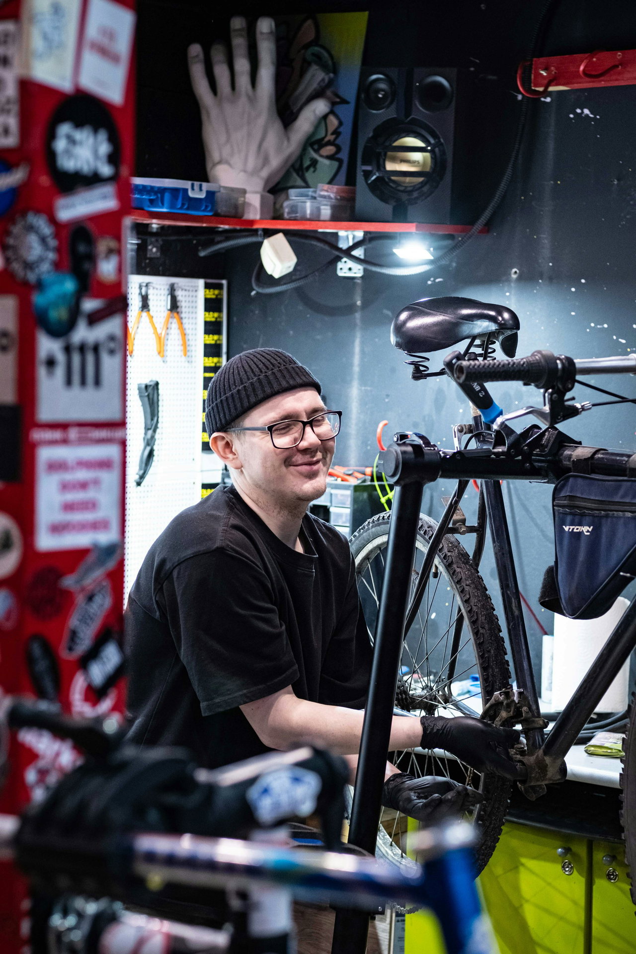 Lächelnder Mechaniker mit Mütze und Brille arbeitet in einer bunten Werkstatt an einem Fahrrad im Ständer