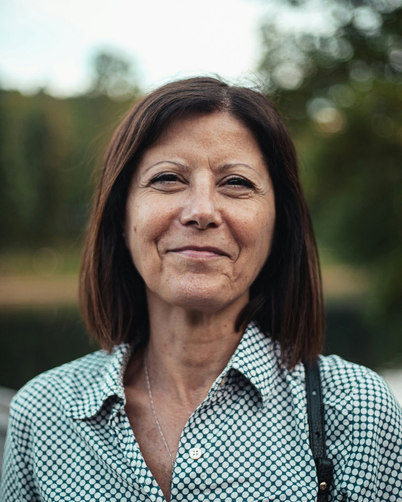 Lächelnde Frau mit dunkelbraunem Bob und kariertem Hemd, im Freien vor unscharfem Wald und See