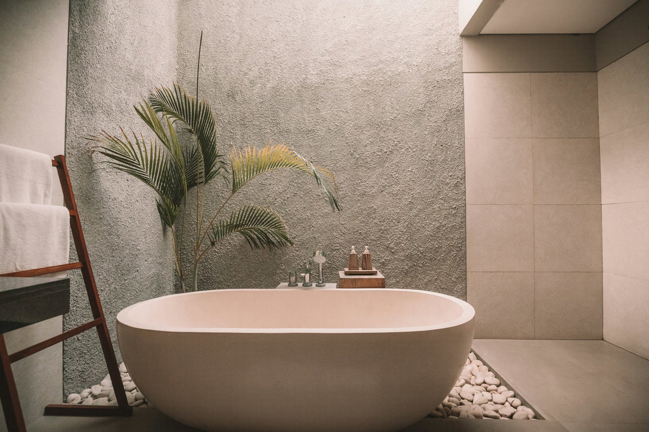 Freistehende helle Badewanne vor einer grauen Wand aus grobem Kalkputz, daneben eine Palme und eine Handtuchleiter aus Holz, am Boden weiße Kiesel.