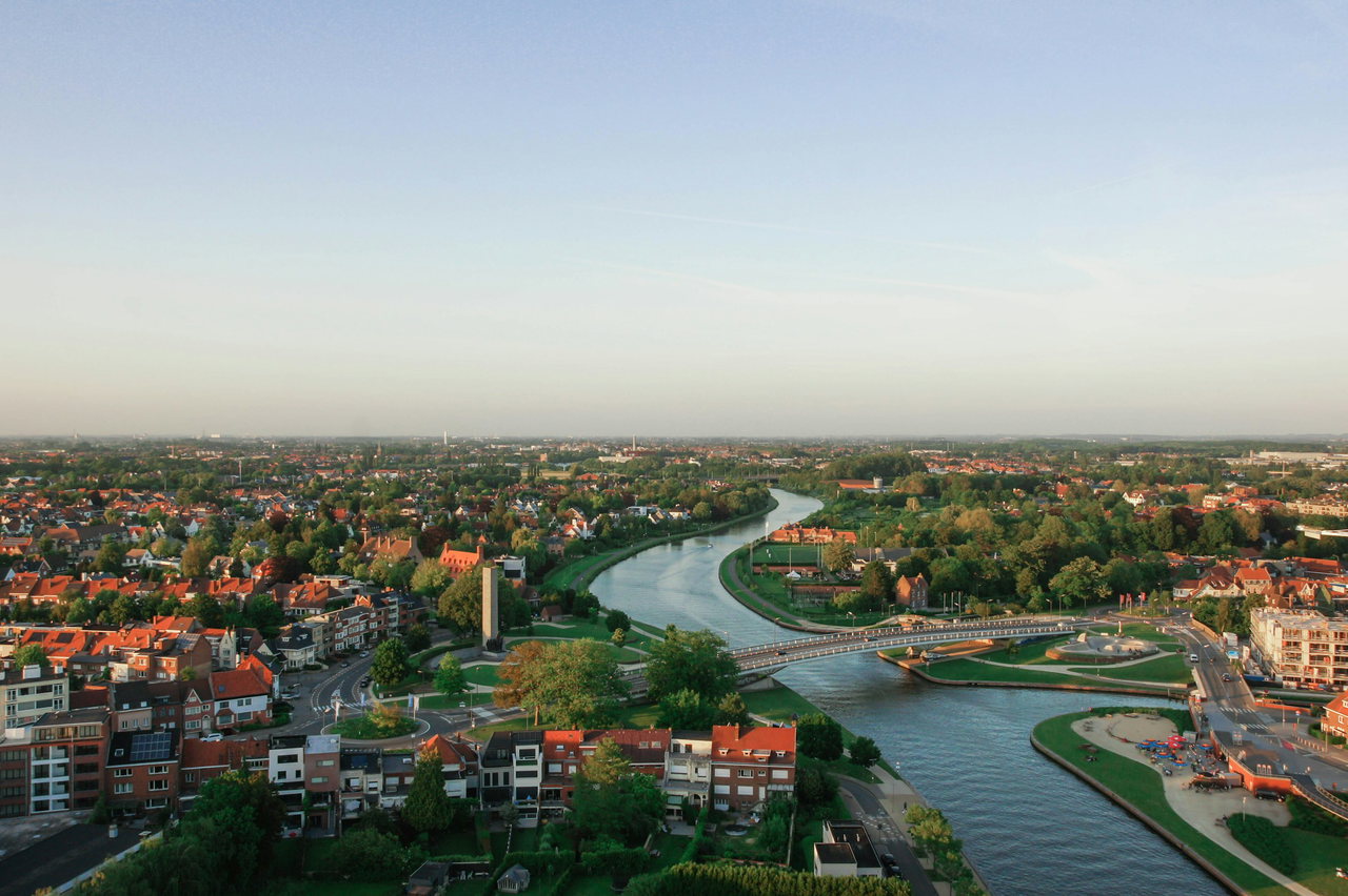 Luftaufnahme eines Stadtviertels mit roten Dächern, Grünanlagen und einem geschwungenen Kanal mit Brücke im Abendlicht.