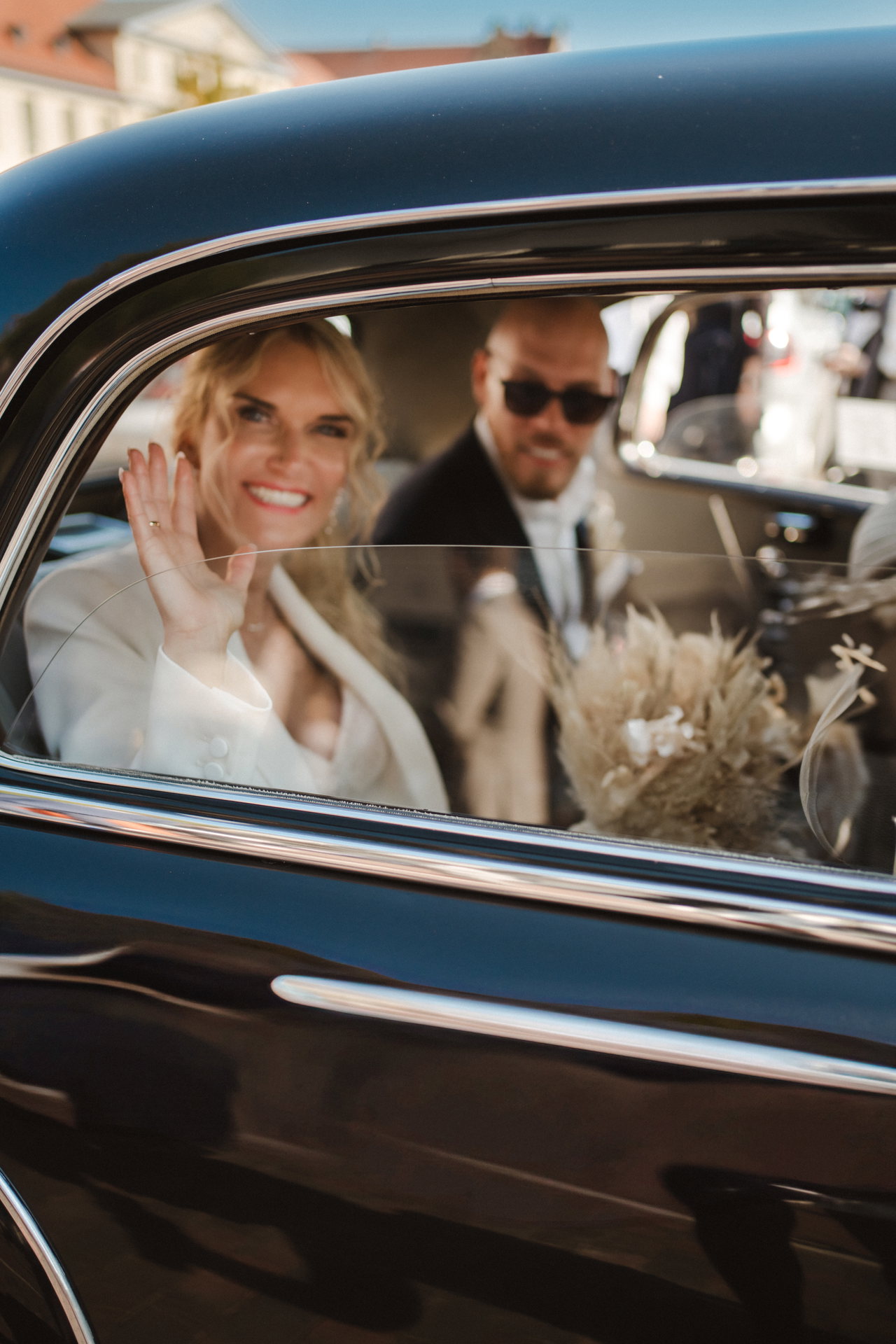 Elain winkt aus dem Autofenster, Marcel mit Sonnenbrille neben ihr
