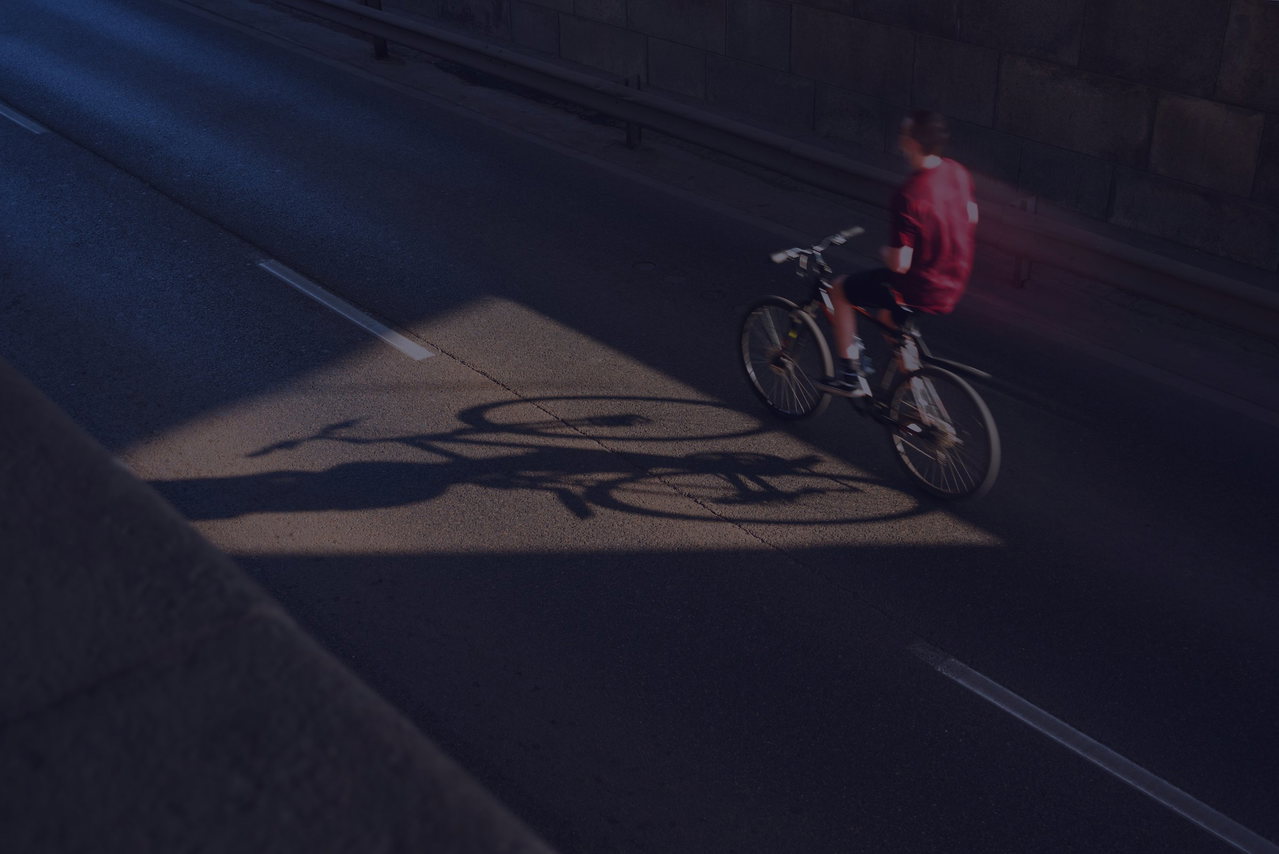 Radfahrer im roten Shirt fährt über eine dunkle Stadtstraße, ein Sonnenfleck wirft seinen langen Schatten auf den Asphalt