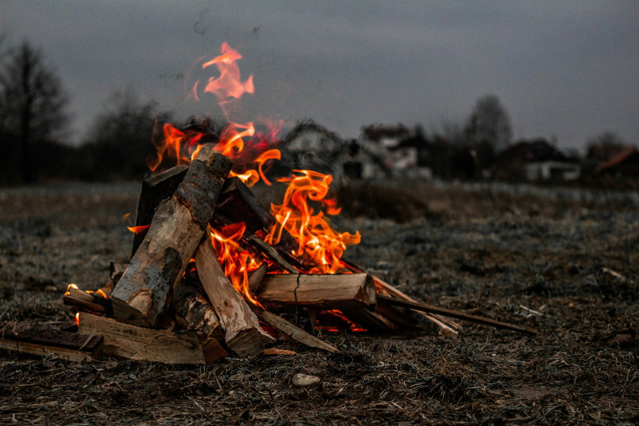 Aufgeschichtetes Holz brennt auf einem Feld in der Dämmerung, im Hintergrund die Häuser eines Dorfes