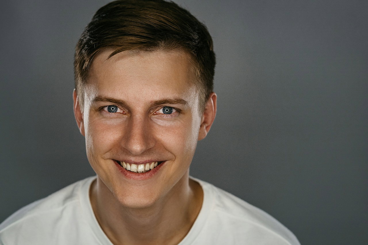 Strahlend lächelnder junger Mann mit kurzen braunen Haaren und weißem Shirt blickt direkt in die Kamera vor grauem Hintergrund