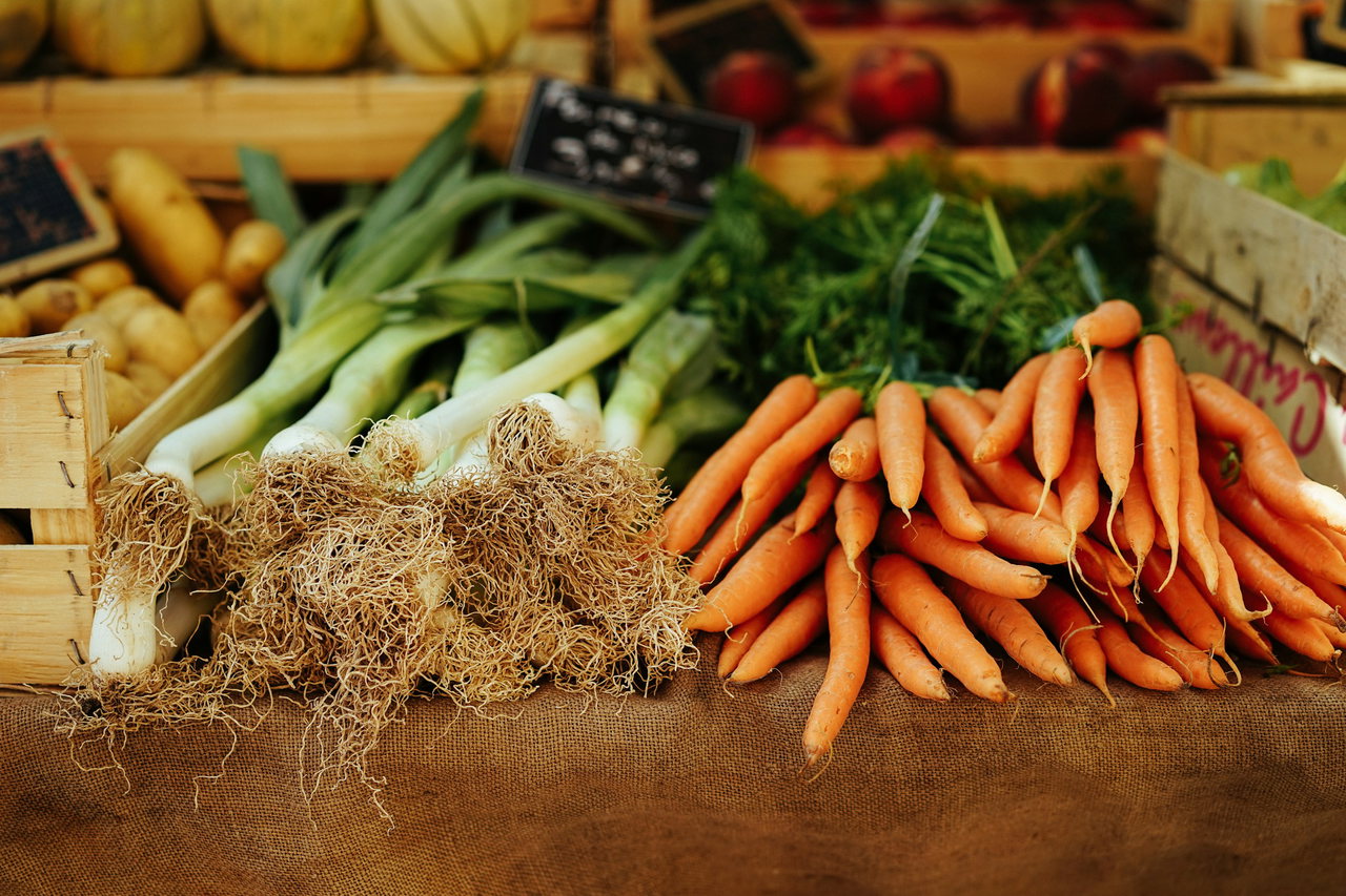 Marktstand mit Bündeln frischer Möhren und Lauch auf Jute, dahinter Holzkisten mit Kartoffeln und rotem Obst