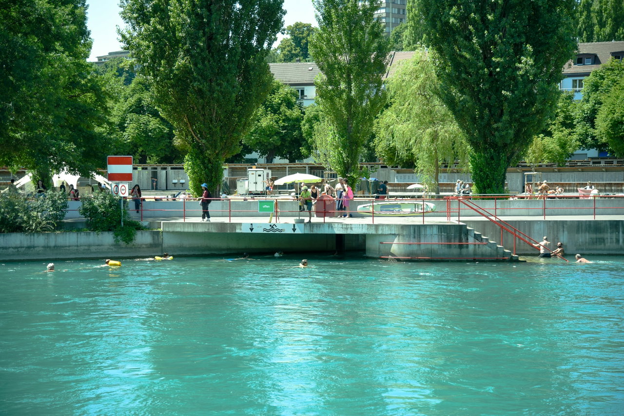 Türkisblaues Flussbad im Sommer mit Badegästen, Liegewiese und hohen Pappeln am Ufer
