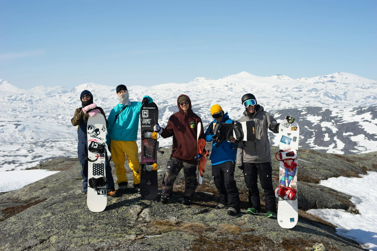 Gruppe von Snowboardern und Skifahrern posiert mit Brettern auf einem Felsen vor verschneiten Bergen