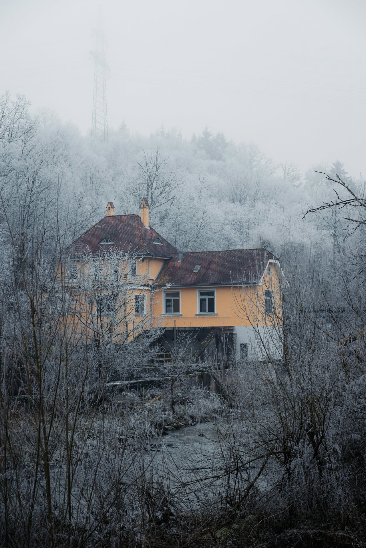 Gelbes Landhaus am Hang im Raureif, umgeben von frostigen Bäumen und aufsteigendem Winternebel