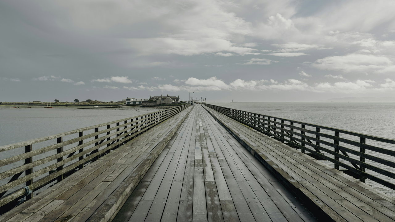 Lange hölzerne Seebrücke mit Geländern über ruhigem Wasser, am Horizont flaches Land mit Häusern, darüber Wolken