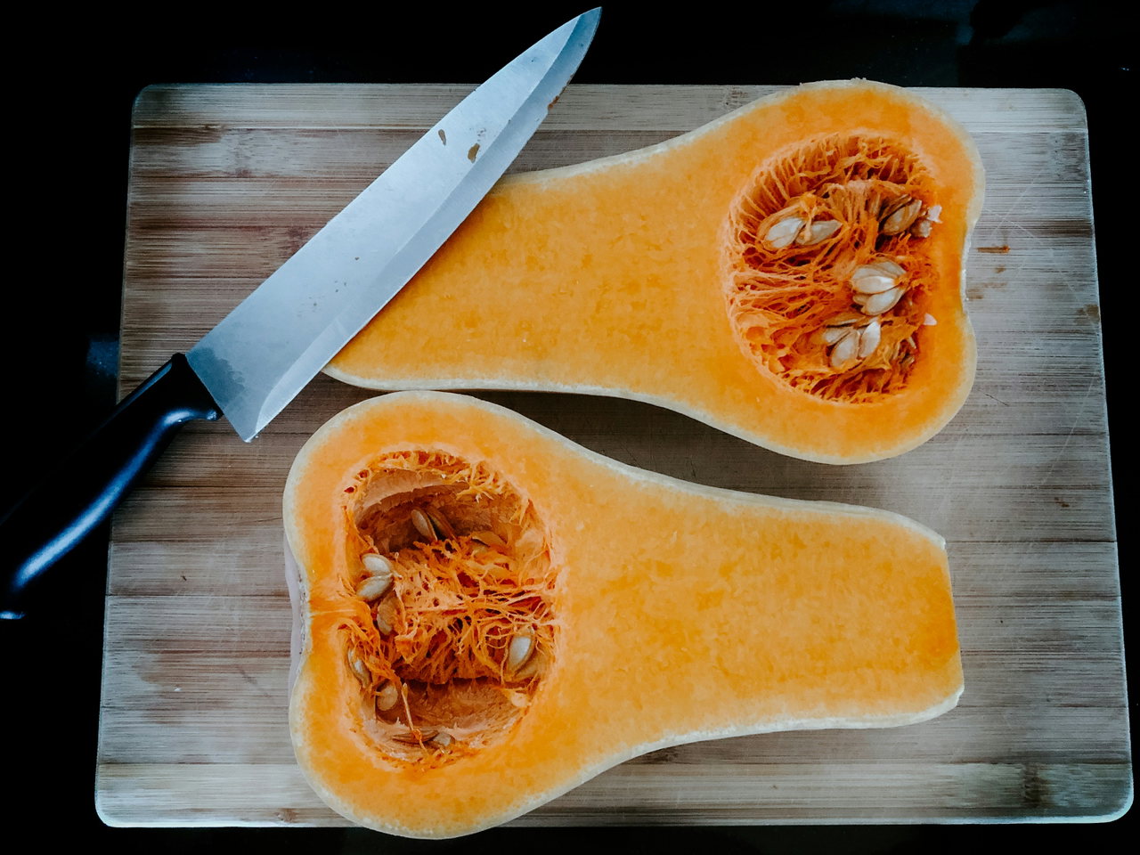 Halbierter Butternut-Kürbis mit Kernen auf einem Holzbrett neben einem Küchenmesser