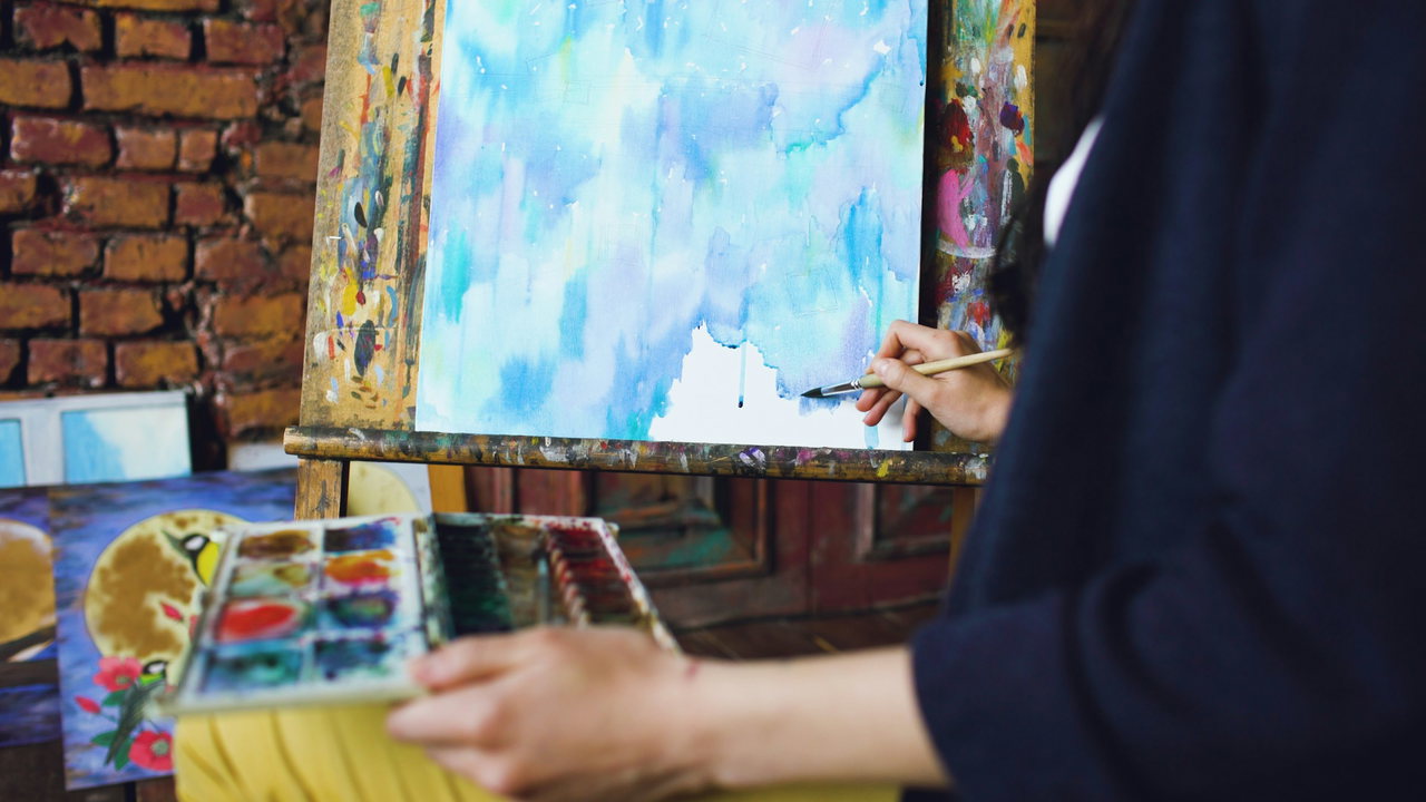 Eine Hand malt mit dem Pinsel ein Aquarell an der Staffelei, daneben Farbkasten und Backsteinwand.