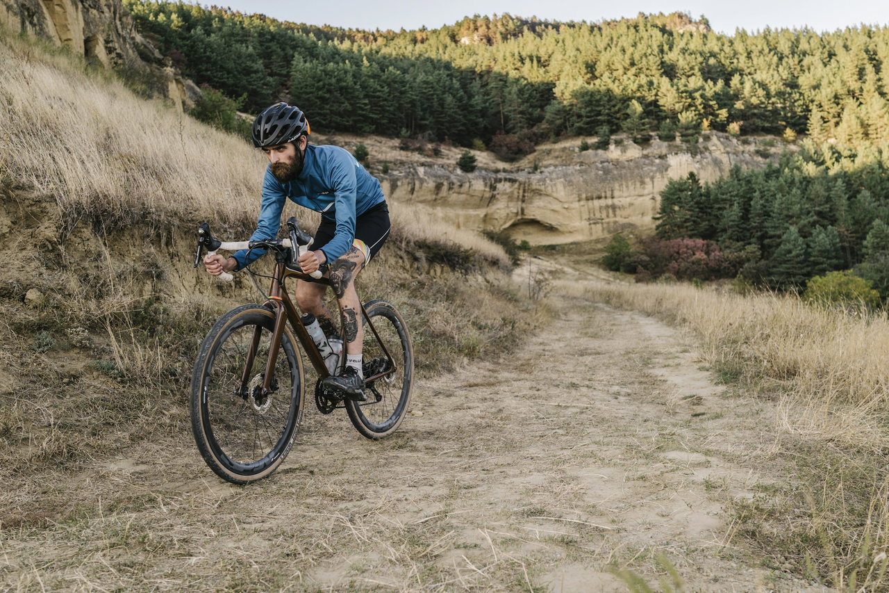Bärtiger Radfahrer mit Helm fährt auf einem Gravelbike über einen sandigen Schotterweg zwischen Kiefernhängen
