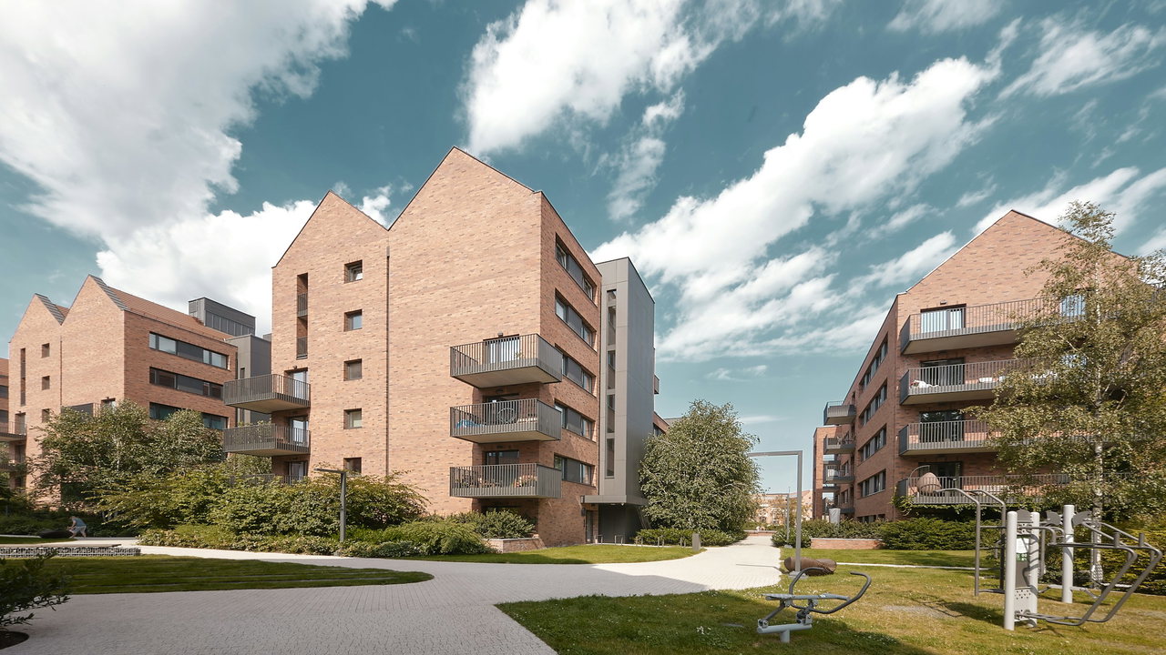 Innenhof zwischen Wohnhäusern aus hellrotem Klinker mit Spitzgiebeln und Balkonen, mit Rasenflächen, gepflasterten Wegen, Bäumen und Fitnessgeräten.