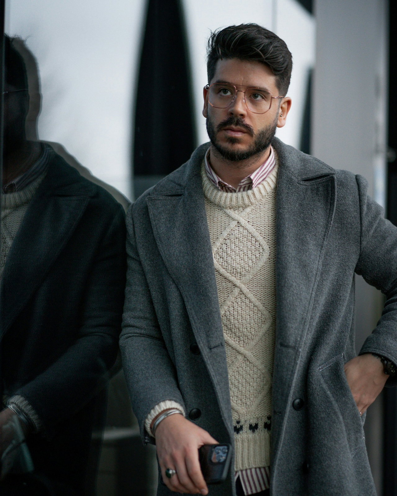 Mann mit Brille trägt grauen Wollmantel über cremefarbenem Zopfstrickpullover und gestreiftem Hemdkragen