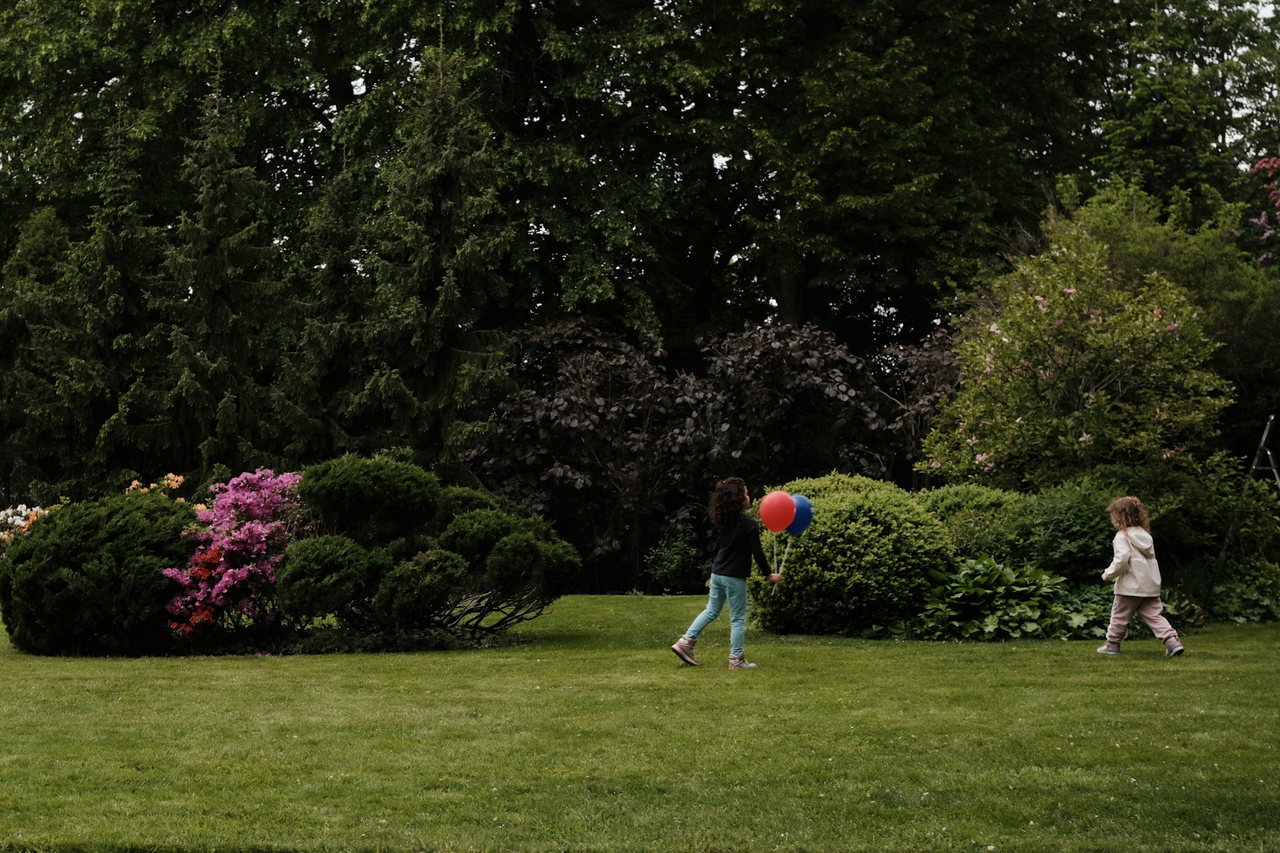 Zwei kleine Kinder spielen auf einer gepflegten Wiese im Park, eines hält rote und blaue Luftballons.