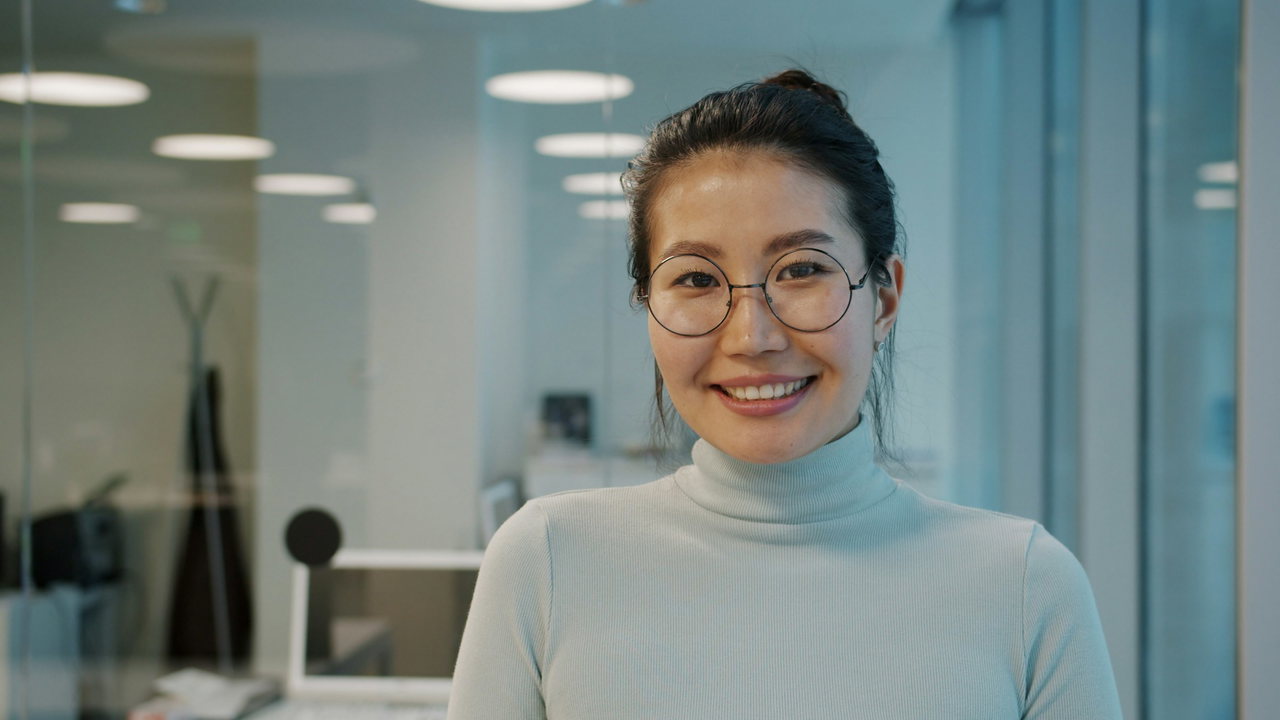 Lächelnde Frau mit Rundbrille und hellem Rollkragenpulli in einem modernen, hellen Büro mit Glaswänden