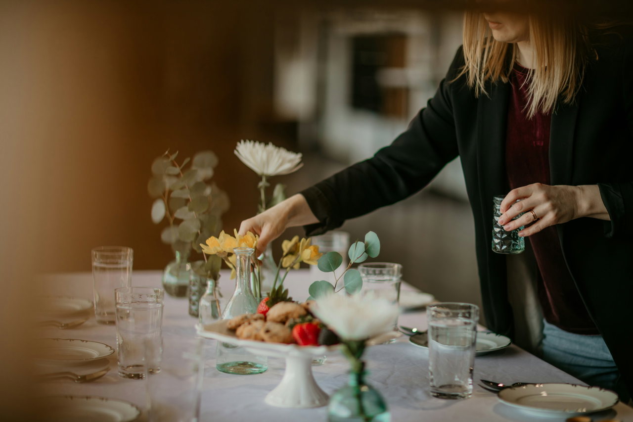 Servicekraft ordnet an einer festlich gedeckten Tafel Blumen und Gläser, warmes Licht
