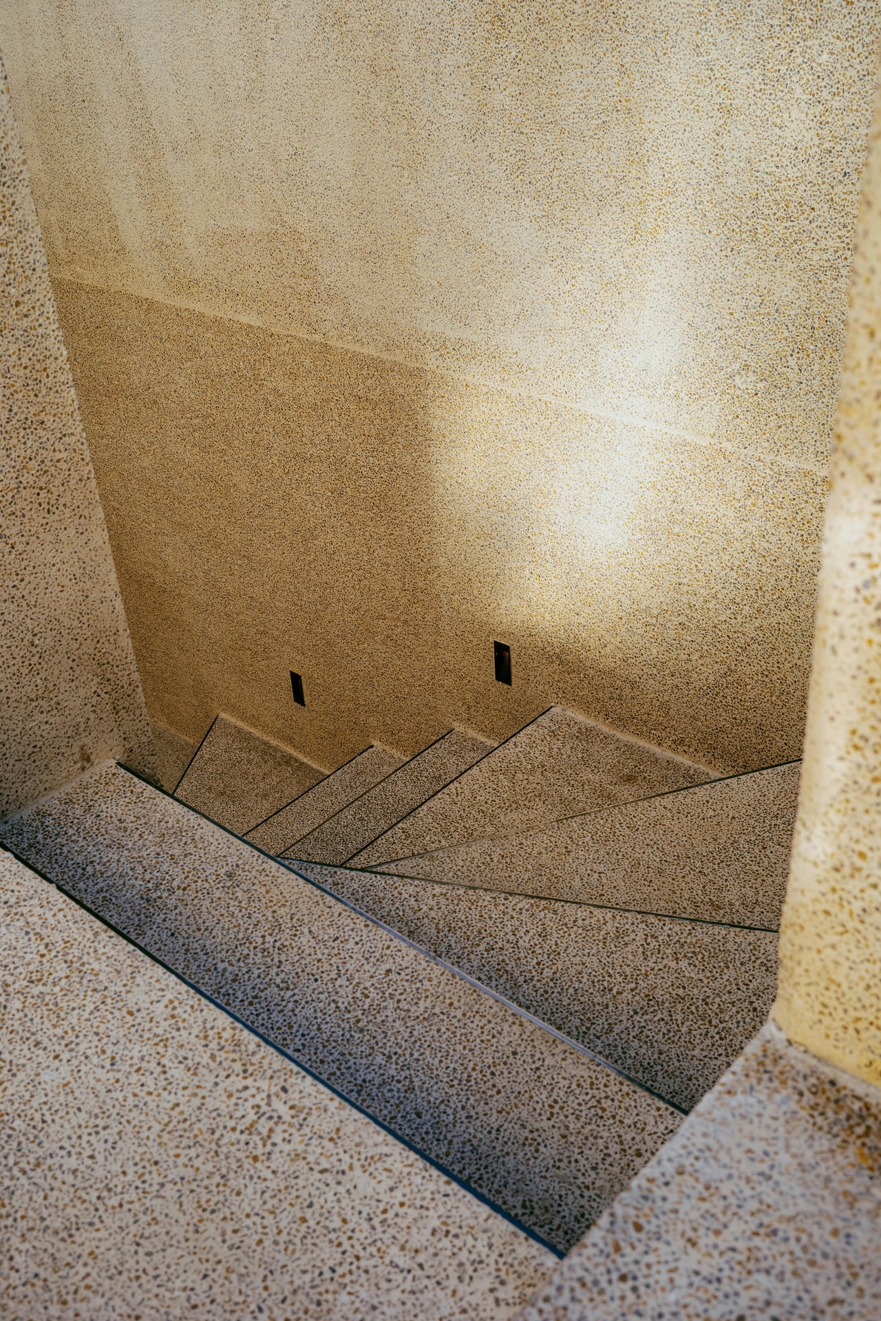 Blick von oben in ein Treppenhaus mit gewendelter Treppe aus hellem Terrazzo, auf die Wand fällt warmes Licht.
