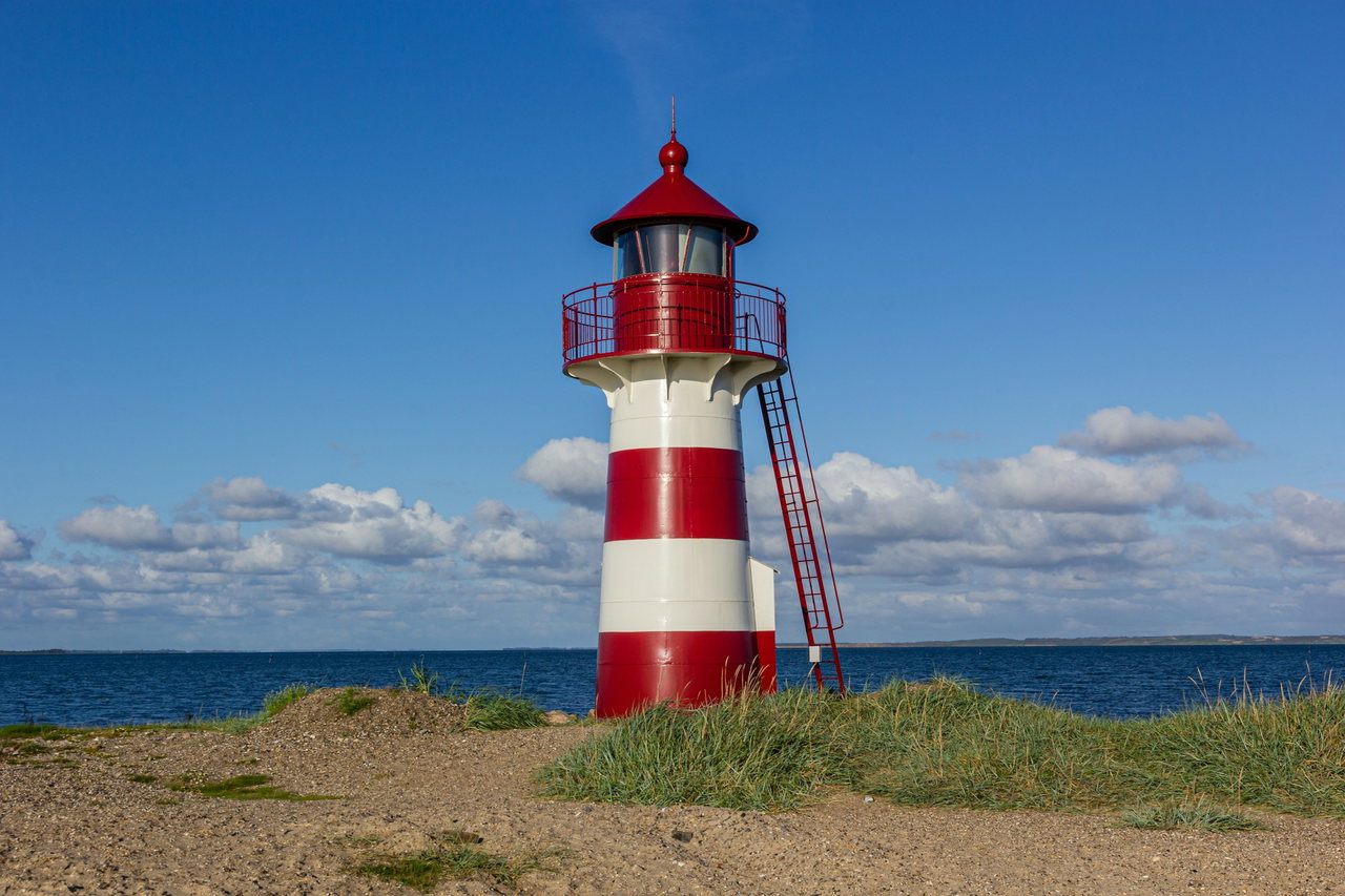 Kleiner rot-weiß gestreifter Leuchtturm mit roter Laterne und Leiter auf grasbewachsenem Ufer vor tiefblauer See