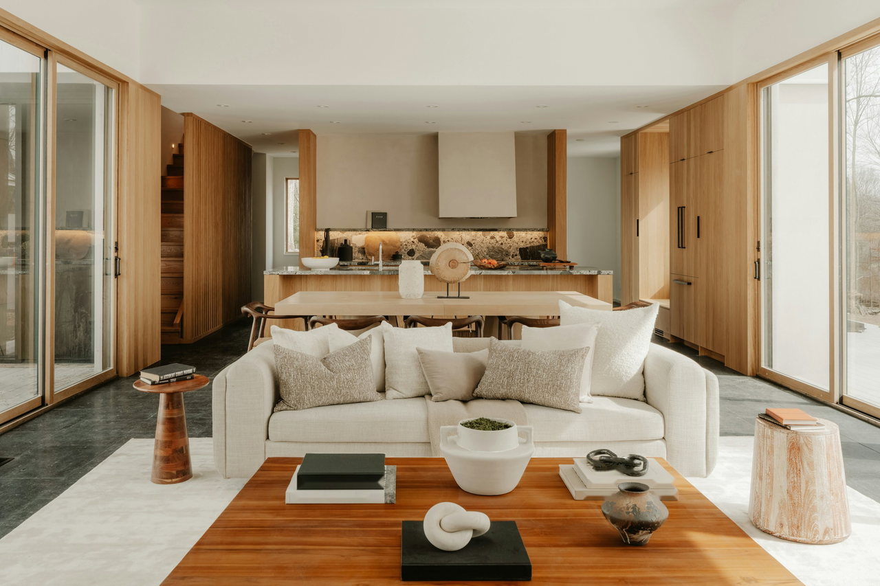 Offener Wohnraum mit hellem Sofa und Couchtisch aus Holz zwischen raumhohen Holzeinbauten und Glasschiebetüren, dahinter Essplatz und Küche mit Steinrückwand.