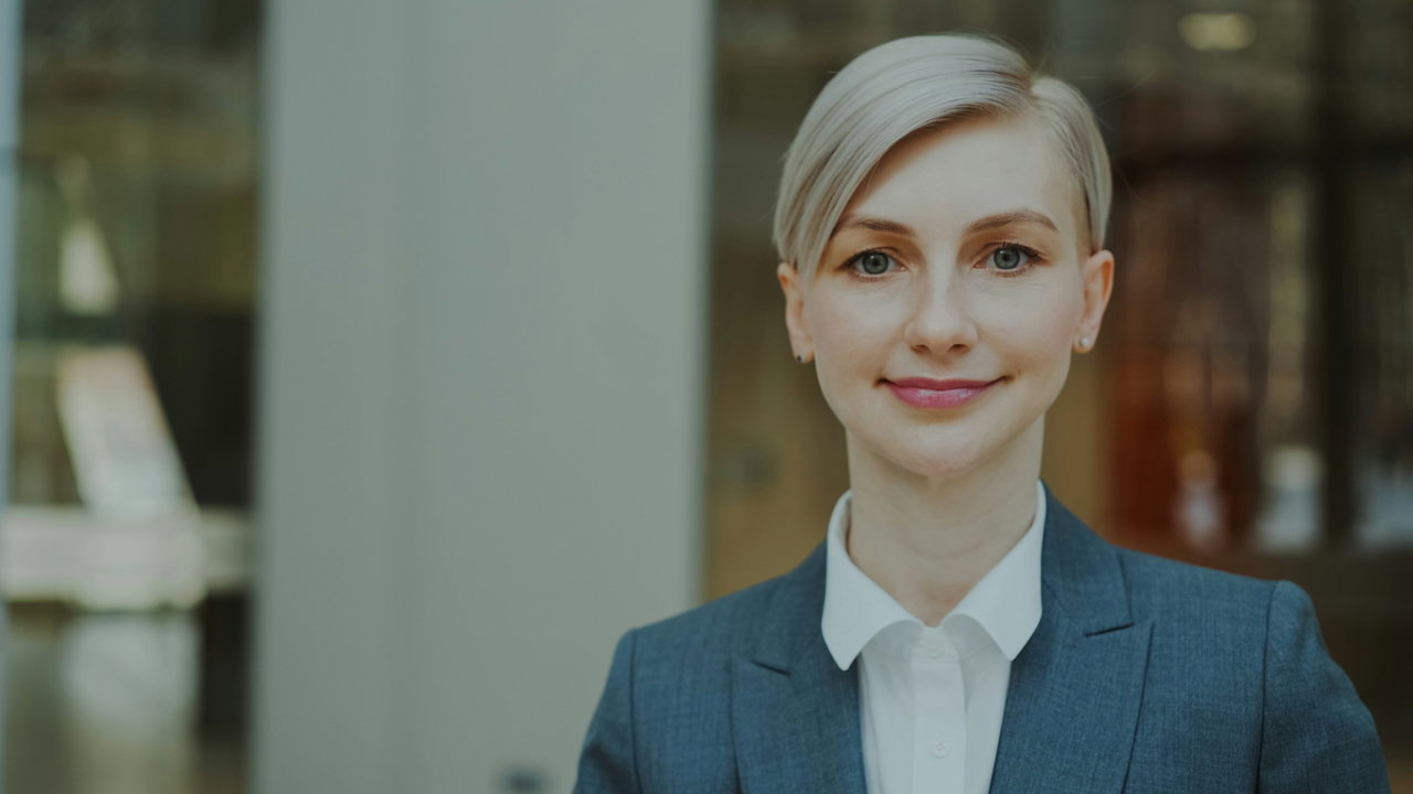 Porträt einer Frau mit kurzem hellblondem Haar im blauen Blazer und weißer Bluse vor unscharfem Bürohintergrund