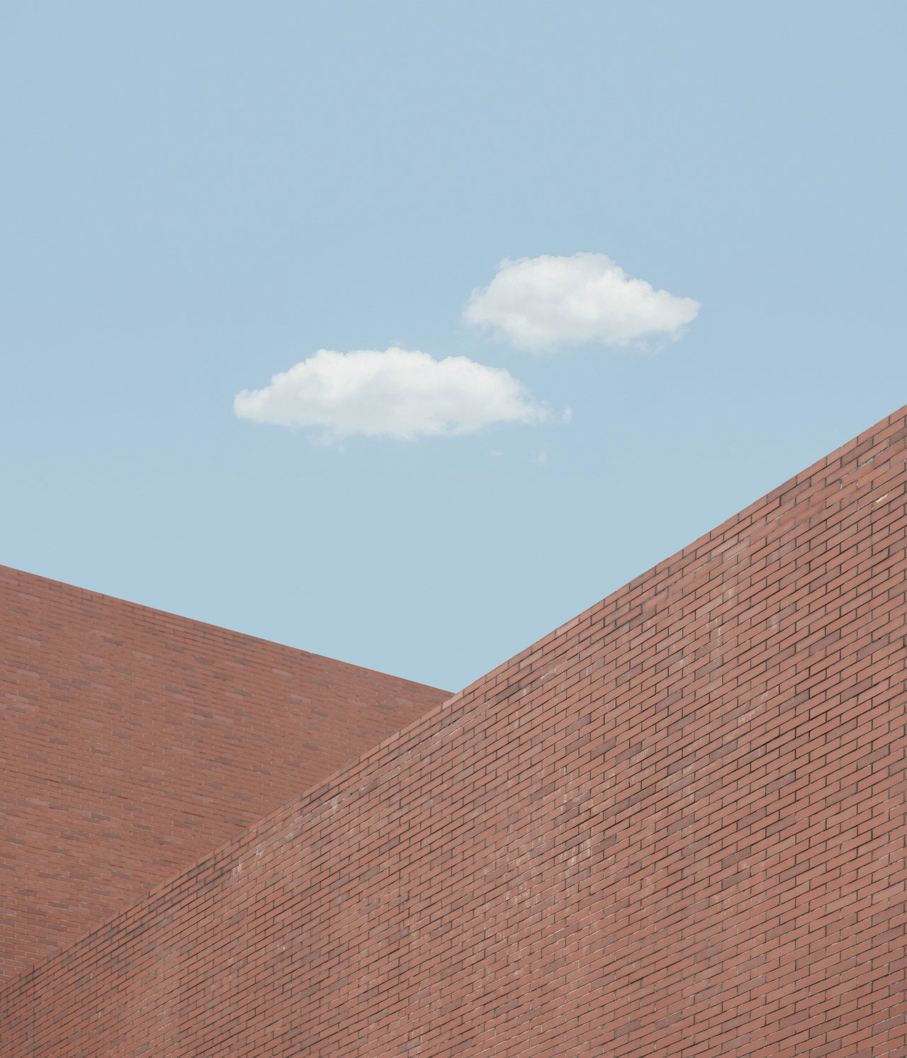 Zwei schräg ansteigende Wandflächen aus rotem Klinker vor hellblauem Himmel mit zwei kleinen Wolken.
