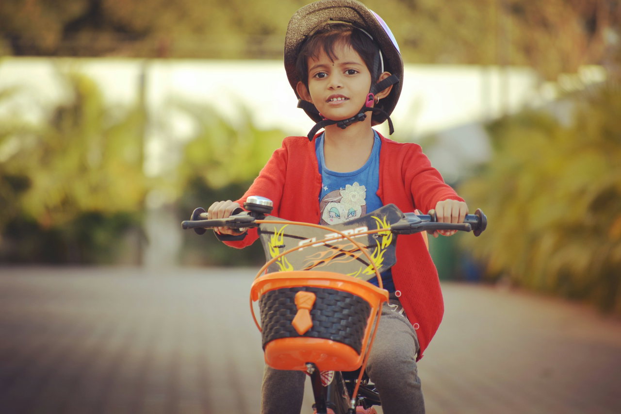 Kind mit Fahrradhelm und roter Jacke fährt auf einem Kinderrad mit orangefarbenem Lenkerkorb auf die Kamera zu