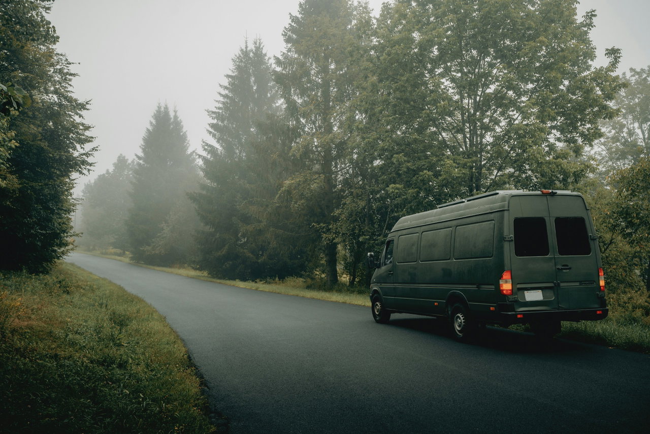 Dunkelgrüner Lieferwagen ohne Beschriftung parkt an einer nebligen Landstraße zwischen Nadelbäumen