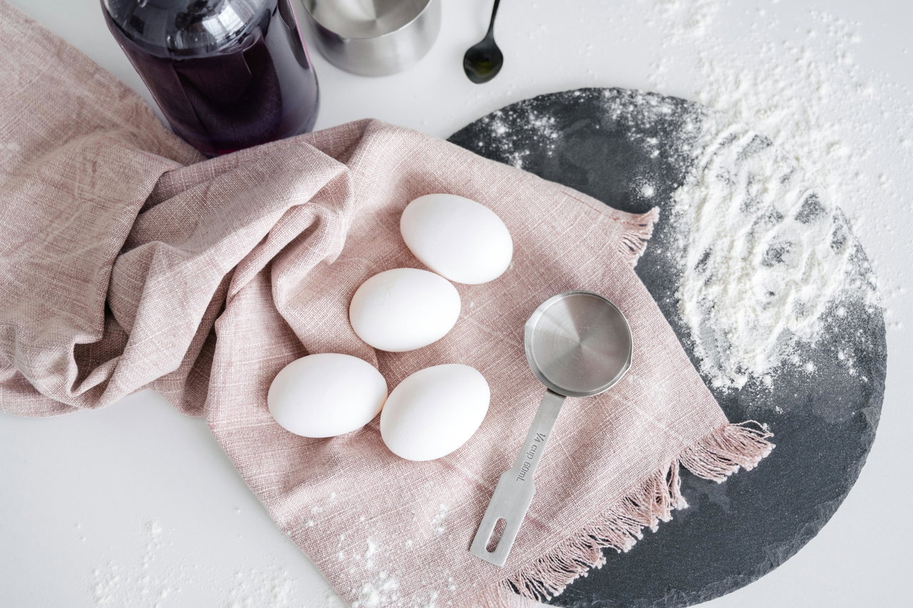 Eier, ein Messlöffel und Mehl auf einem rosafarbenen Tuch