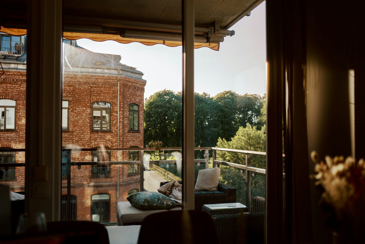 Blick aus einem Wohnraum durch die offene Glastür auf eine sonnige Loggia mit Kissen und Markise, gegenüber ein Backsteinbau und hohe Bäume im Abendlicht.